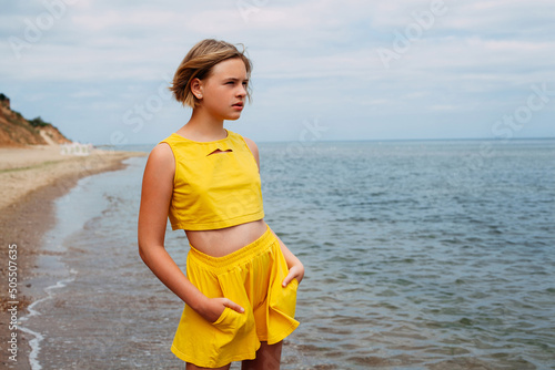 A portrait of a young tanned girl in yellow summer clothes stands on the seashore against the backdrop of sandy hills  holds her hands in her pockets and looks into the distance.