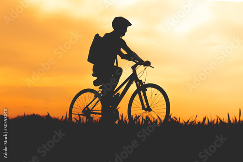 Silhouette of a cyclist at sunset with a helmet and a backpack on his way through the countryside. © Yuri Hoyda