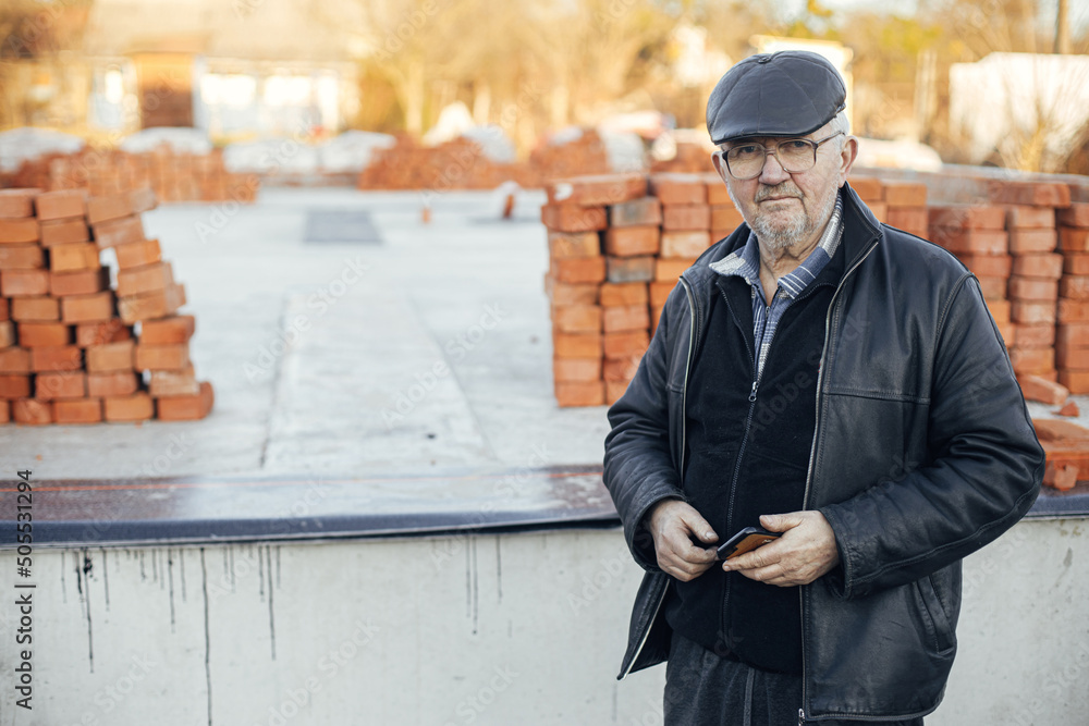 Senior handyman standing with phone on background of bricks masonry with cement on concrete foundation. Foreman and process of house building at construction site Senior handyman standing with phone on background of bricks masonry with cement on concrete foundation. Foreman and process of house building at construction site