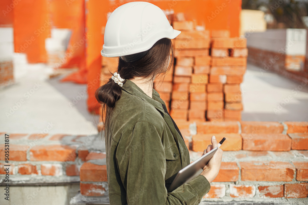Stylish woman architect with tablet blueprints at construction site. Young female engineer or construction worker in hardhat looking at plans of new modern house