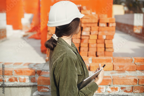 Stylish woman architect with tablet blueprints at construction site. Young female engineer or construction worker in hardhat looking at plans of new modern house © sonyachny