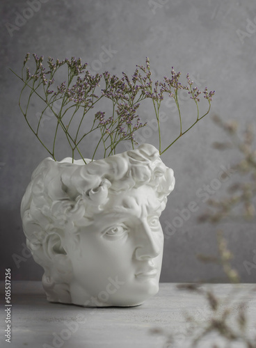 Close-up of a white plaster statue of the head of David on a gray concrete background. Minimalist artistic flower poster. Ancient Greek sculpture.