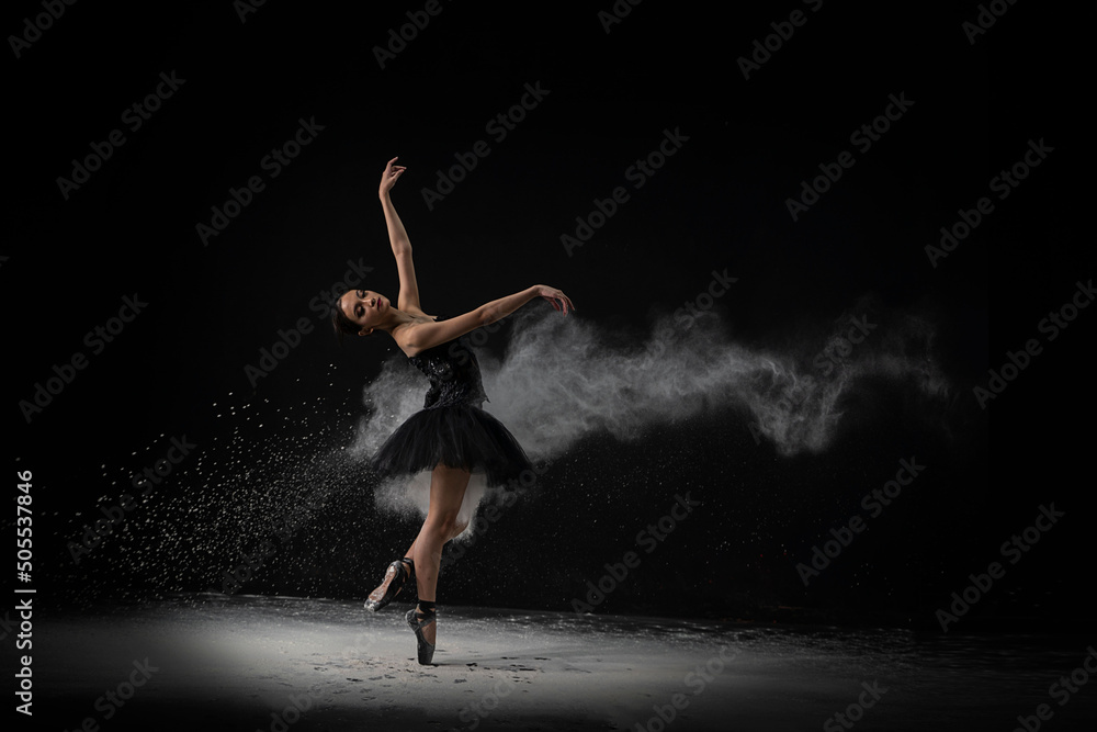 Emotional Southeast Asian ballet dancer in a black dress performing a move on a black background - obrazy, fototapety, plakaty 