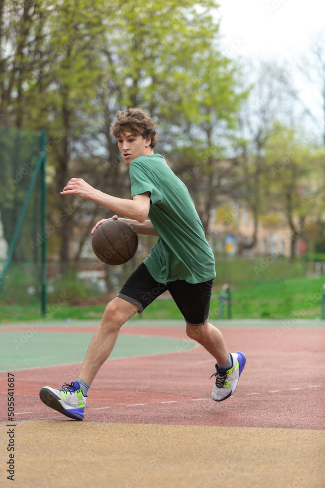 Cute young teenager in green t shirt with a ball plays basketball on court. Sports, hobby, active lifestyle for boys