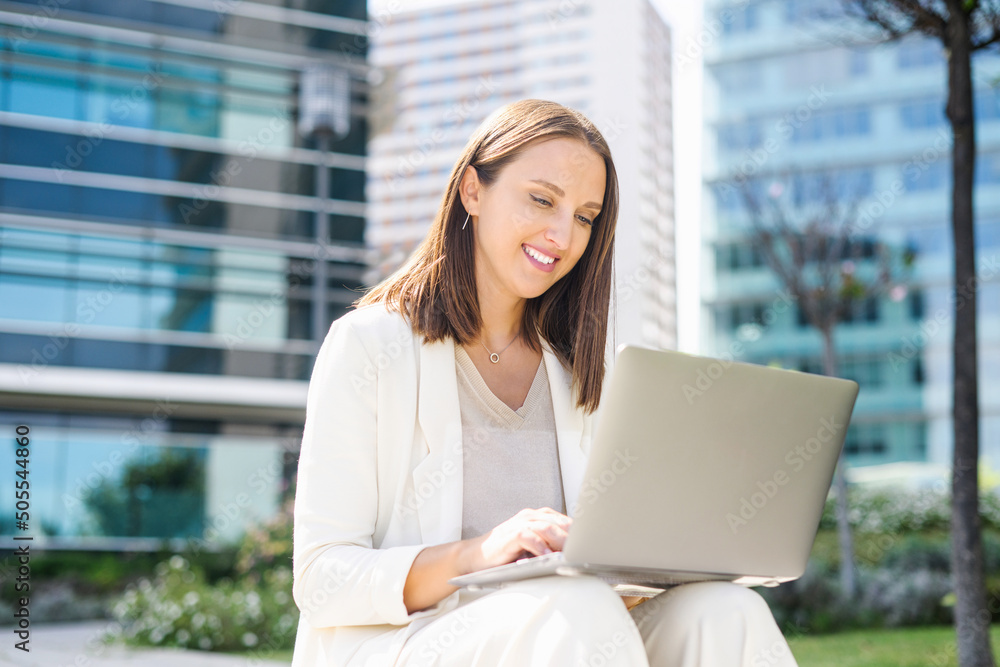 Carefree female office employee typing on the laptop sitting on the step in the city, businesswoman or freelancer woman responds to emails, working outdoors, urban landscape on the background Carefree female office employee typing on the laptop sitting on the step in the city, businesswoman or freelancer woman responds to emails, working outdoors, urban landscape on the background
