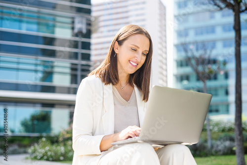 Carefree female office employee typing on the laptop sitting on the step in the city, businesswoman or freelancer woman responds to emails, working outdoors, urban landscape on the background