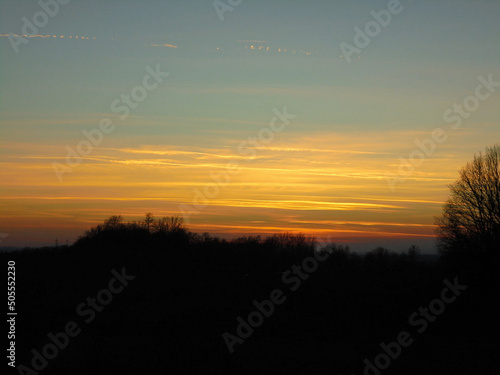 Abstract image: various layers of orange clouds during sunset ranging from blue sky to red cloud at the horizon covered by bare trees.