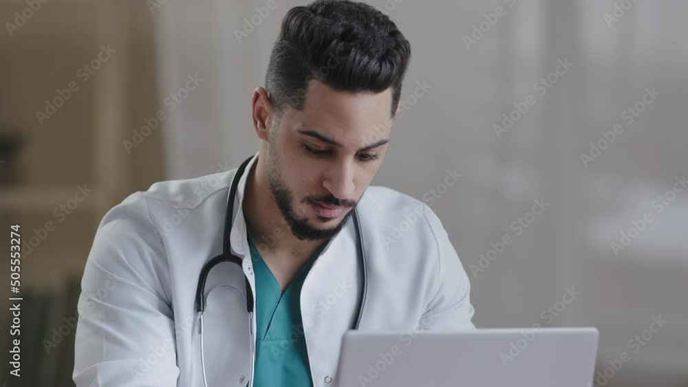 Concentrated man hispanic doctor physician wear white coat use computer internet at desk type recipe for patient on computer at hospital cabinet male young intern consult distantly online medical chat Concentrated man hispanic doctor physician wear white coat use computer internet at desk type recipe for patient on computer at hospital cabinet male young intern consult distantly online medical chat