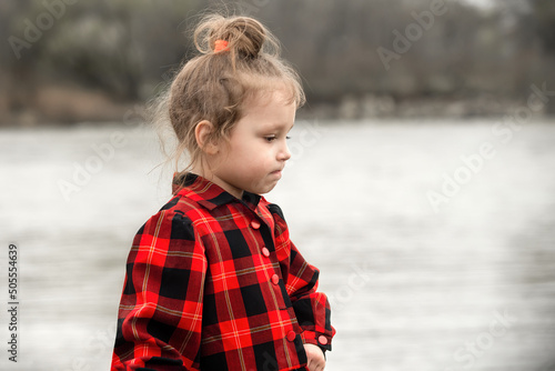 portrait of a little girl with blond hair in a bright red checkered dress against the backdrop of nature