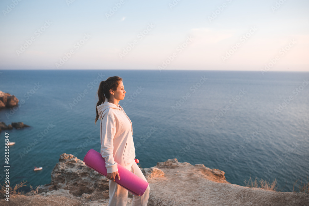 Happy smiling woman holding pink yoga mat wear sportswear hoodie and pants standing on top of rock over sea side background. Pretty adult girl practice fitness at open air outside. Healthy lifestyle.