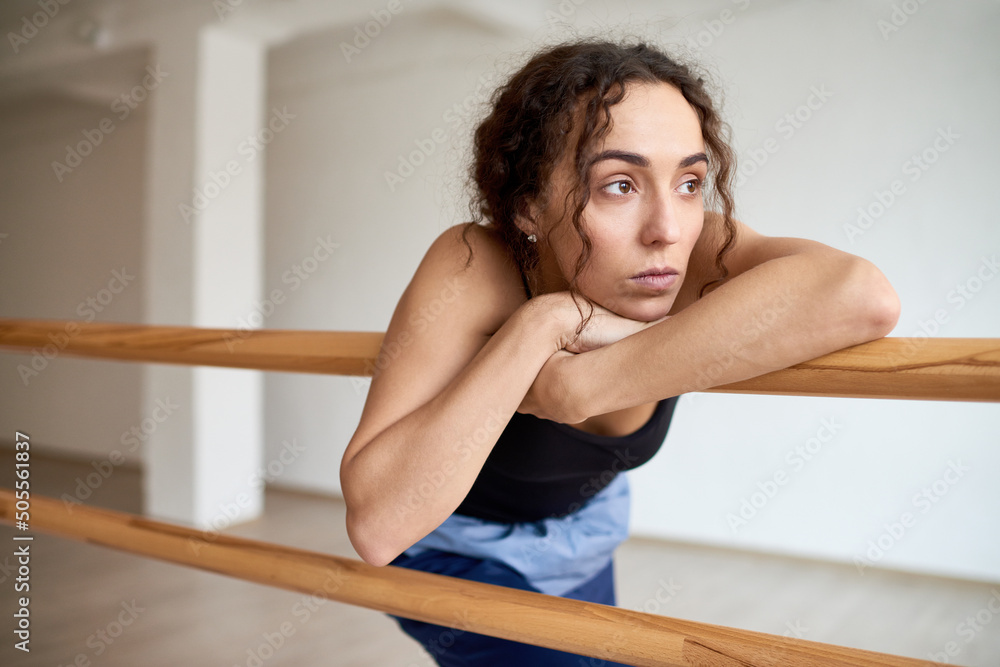 Young attractive curly woman looking into the distance thoughtfully while leaning on ballet barre in dancing hall Young attractive curly woman looking into the distance thoughtfully while leaning on ballet barre in dancing hall