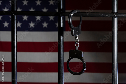 Police handcuffs are fastened to the bars of the prison cell against the background of the American flag, selective focus.