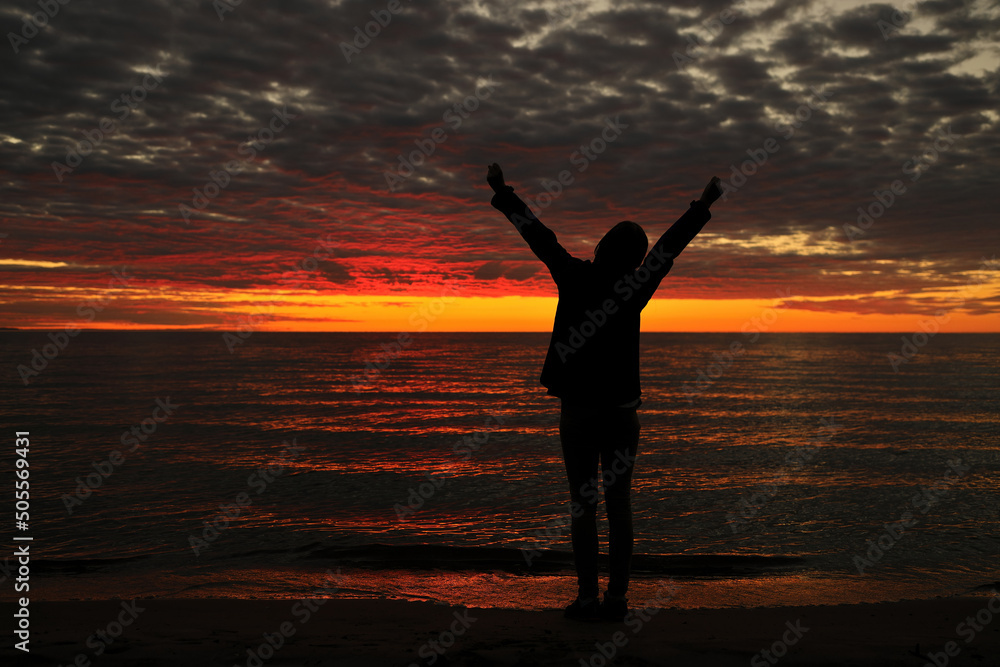 Happy woman sihouette with arms raised up in success to sunset glow at beach by water