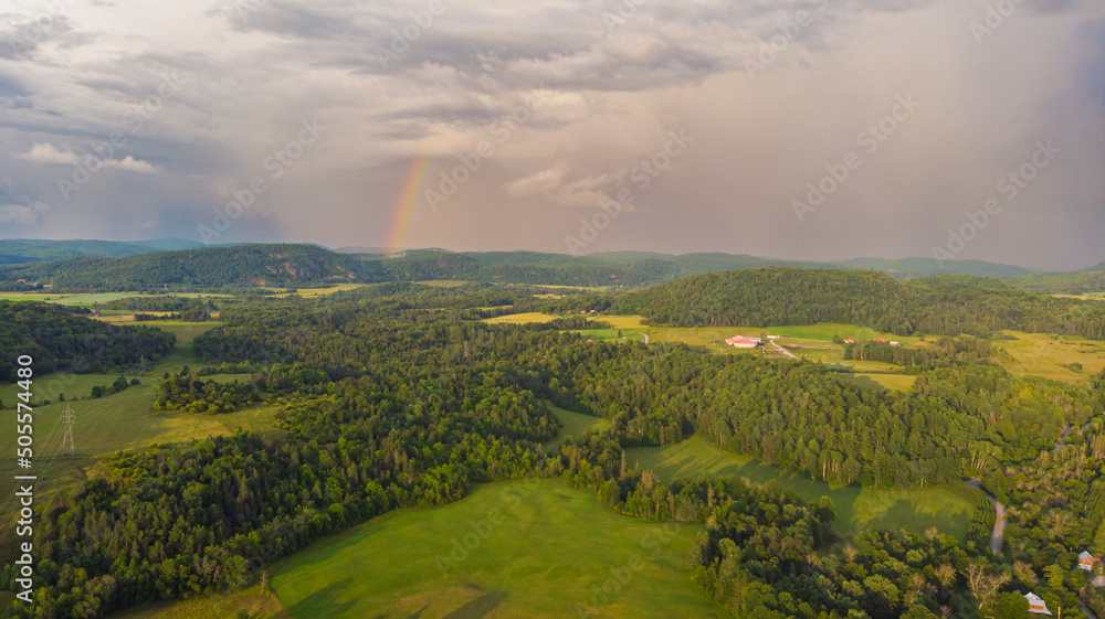 Rainbow over the wide endless horizon of Canada landscape. Green meadow, lush forest on hilly landscape. Rain clouds against blue sky, sunbeams shine through dark clouds. Rainbow rises on the horizon