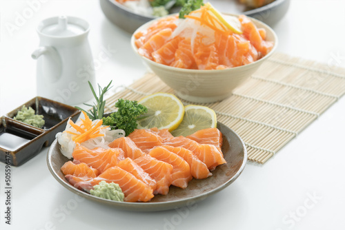 Selective focus on sliced salmon sashimi in plate served with wasabi, soy sauce, sliced lemon, carrot and radish at kitchen. Salmon on ice in bowl on blur background. Japanese food home cooked concept