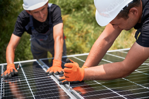 Male workers building photovoltaic solar panel system outdoors. Men engineers placing solar module on metal rails, wearing construction helmets and work gloves. Renewable and ecological energy.