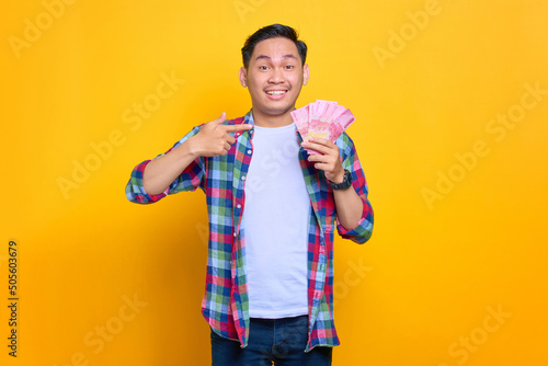 Excited young Asian man in plaid shirt pointing fingers at money banknotes isolated on yellow background