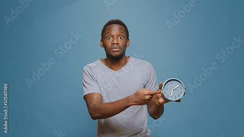 Stressed man checking time on wall clock and running late to work, looking at hour and minutes. Young adult in delay, doing alarm countdown to measure time, being impatient and punctual.