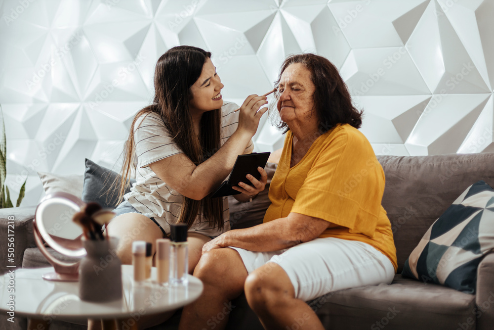 Neta adulta aplicando maquiagem em sua avó. Avó e neta se divertindo em casa. - obrazy, fototapety, plakaty 