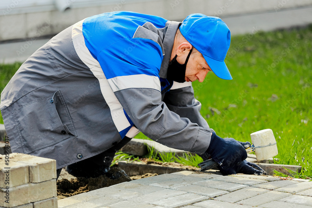 Bricklayer lays paving slabs outside. Working man performs landscaping. Builder lays out sidewalk with stone blocks. Authentic workflow. Hard work in old age. Bricklayer lays paving slabs outside. Working man performs landscaping. Builder lays out sidewalk with stone blocks. Authentic workflow. Hard work in old age.