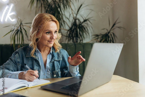 Beautiful mature businesswoman using laptop and writing something to notebook in cafe