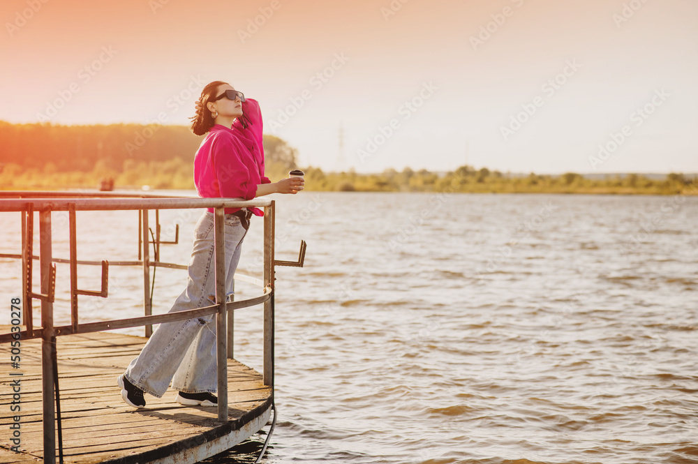 Brunette woman standing on the edge of pier on the lakeside under bright evening sun