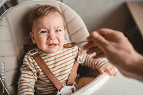 Toddler crying at lunch time sitting in a highchair in the living room at home. Little baby girl refuses to eat off spoon photo