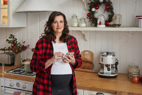 beautiful happy pregnant woman in the kitchen decorated for christmas with a smartphone in her hands. Merry Christmas greetings to loved ones.