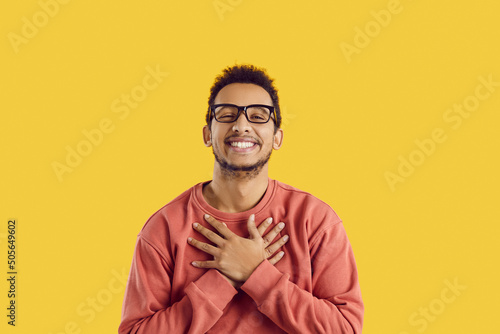 Happy black man in glasses smiling and holding his hands on his chest to express endless gratitude and appreciation. Cheerful ethnic guy says thank you from the bottom of his heart for gift or present