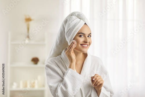 Fotografia Beautiful woman in white bathrobe and towel turban on head looking at reflection in mirror and putting toner on face with cotton disk pad preparing herself for new day