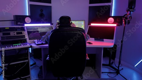 Soundwoman in cap and headphones working in the recording studio with computer and mixer creating music. Woman enjoys process and dances sitting on a chair. Back view full shot photo