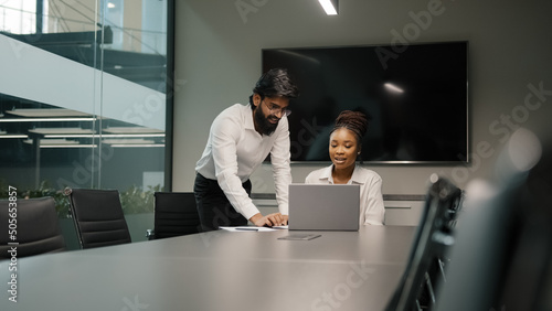 Professional Arabian Indian teacher mentor helping African woman new employee teaching intern explaining online project service corporate app using laptop. Teamwork internet win discussion in office