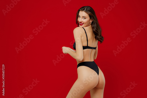 Back side view young sexy hot brunette caucasian woman 20s with perfect fit body wearing black underwear isolated on plain red color background studio portrait. People female beauty lingerie concept