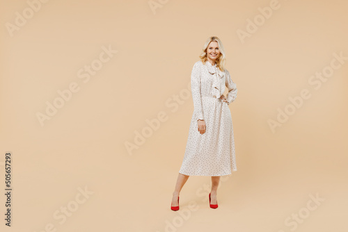 Full size body length charming elderly gray-haired blonde woman lady 40s years old wears pink dress hold hand on waist looking camera posing isolated on plain pastel beige background studio portrait.