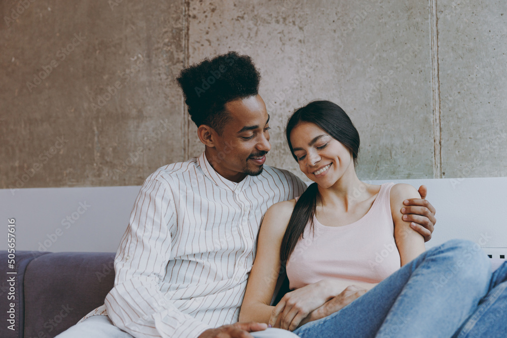 Two young friends smiling happy fun woman man of African American ethnicity 20s in casual clothes hugging sitting on gray sofa indoor spend free time rest in flat together. People lifestyle concept. - obrazy, fototapety, plakaty 