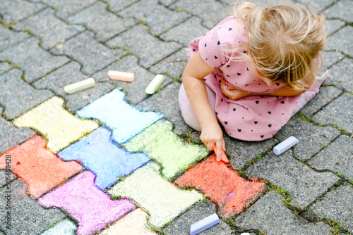 Little preschool girl painting with colorful chalks on ground on backyard. Positive happy toddler child drawing and creating pictures. Creative outdoors activity in summer.