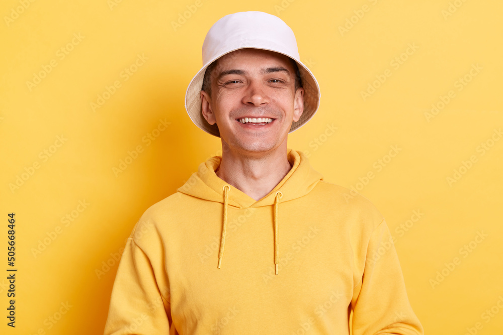 Portrait of satisfied delighted Caucasian man wearing casual style hoodie and panama, looking at camera with positive expression, being happy, posing isolated over yellow background.