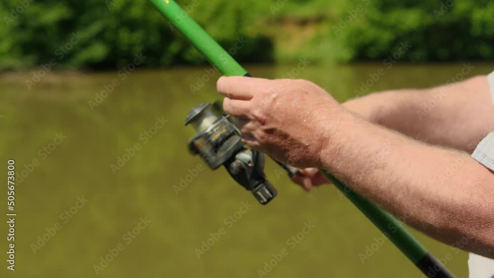 Close-up of a man's hands holding a green fishing rod and turning the reel knobs. In the background, the blurry brown lake water and green foliage. The concept of recreation and hobbies in nature Close-up of a man's hands holding a green fishing rod and turning the reel knobs. In the background, the blurry brown lake water and green foliage. The concept of recreation and hobbies in nature