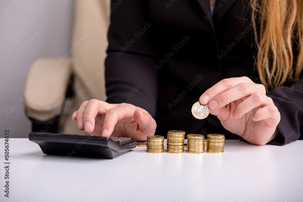 Φωτογραφία The girl is counting on a calculator at the table, in front of stacks of coins,