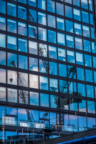Glass facade of a modern office building reflecting the blue sky and two construction cranes