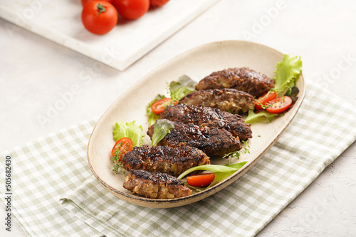 Several homemade roasted minced meat sausages köfte on an oval beige colored plate with green leaves and cherry tomatoes on green checkered kitchen towel