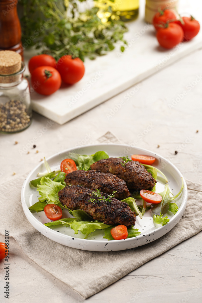Several homemade roasted minced meat sausages köfte on a round white plate with green leaves and cherry tomatoes on beige colored linen napkin