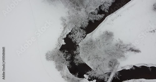 Flying over dark black river and snow covered land. Frosted trees nearby the river, playing and flowy. River is twisting. Winding river flows through winter landscape. Cinematic aerial shot photo