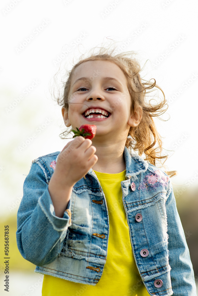 A cute little girl eats strawberries. She is wearing a denim jacket and a yellow T-shirt. Located outside the city. Picnic. Spring. Delicious. Holidays with the family. A cute little girl eats strawberries. She is wearing a denim jacket and a yellow T-shirt. Located outside the city. Picnic. Spring. Delicious. Holidays with the family.
