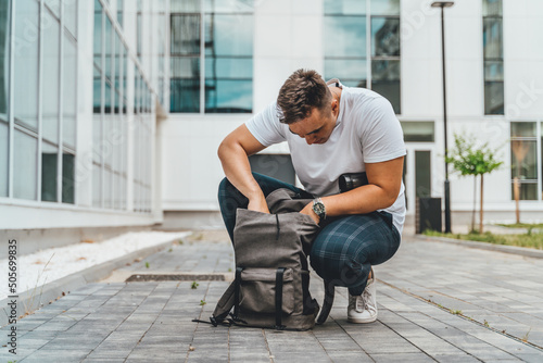 A young student in front of his faculty building searching in his backpack, using phone and tablet and drinking coffee during the day