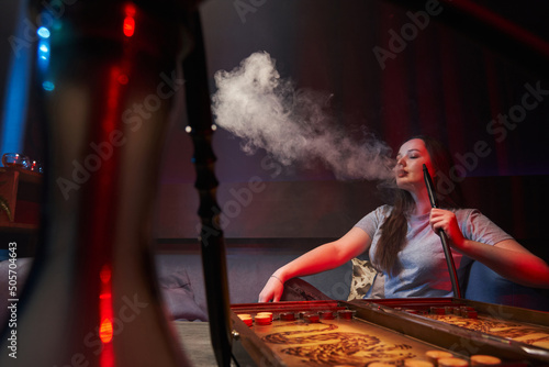 a young woman smokes a hookah, lets out a beautiful cloud of smoke, backgammon is laid out in front of her, a mouthpiece is in her hand