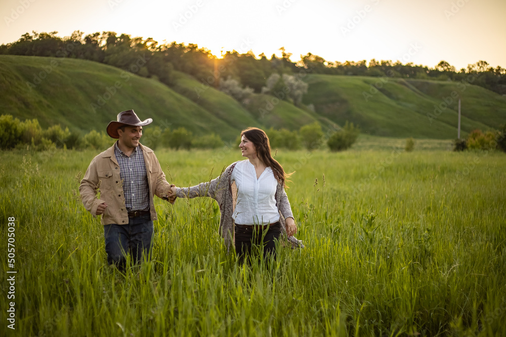 young beautiful couple, on a green meadow in the rays of the setting sun, a man and a girl run towards the camera, cowboy clothes. rustic style