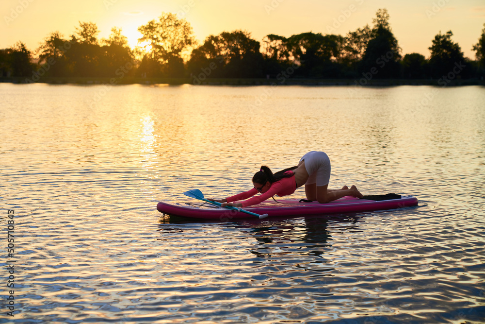 Sporty young woman doing flexible exercise during water activity. Active caucasian brunette practising yoga on sup board during summer sunset.