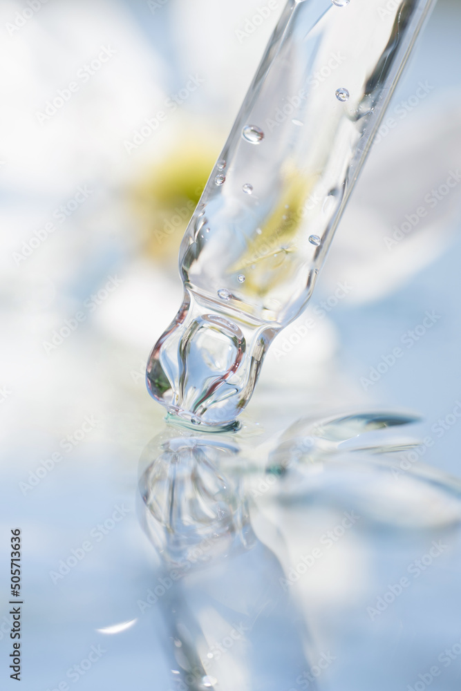 Pipette with transparent cosmetic liquid against white flower on blue background. Texture of face serum or cosmetic oil. Selective focus, vertical image Pipette with transparent cosmetic liquid against white flower on blue background. Texture of face serum or cosmetic oil. Selective focus, vertical image