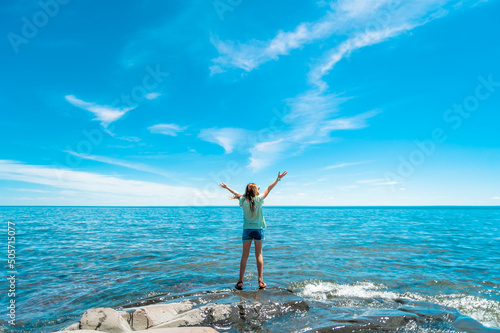 A girl stands on a rocky outcrop in front of a Lake Superior and a bright blue sky with her hands in the air 